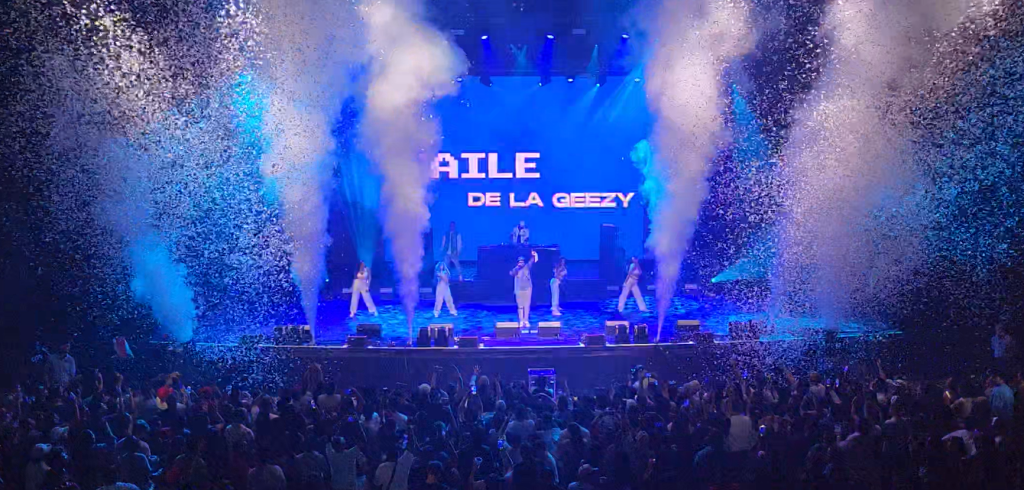 Photo of a concert. On stage there are performers surrounded by smoke. The lighting is blue.