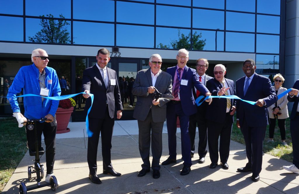 A group of men stand behind a cut blue ribbon, one is holding a large pair of scissors