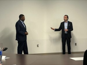 Two men stand in front of a blank wall conversing