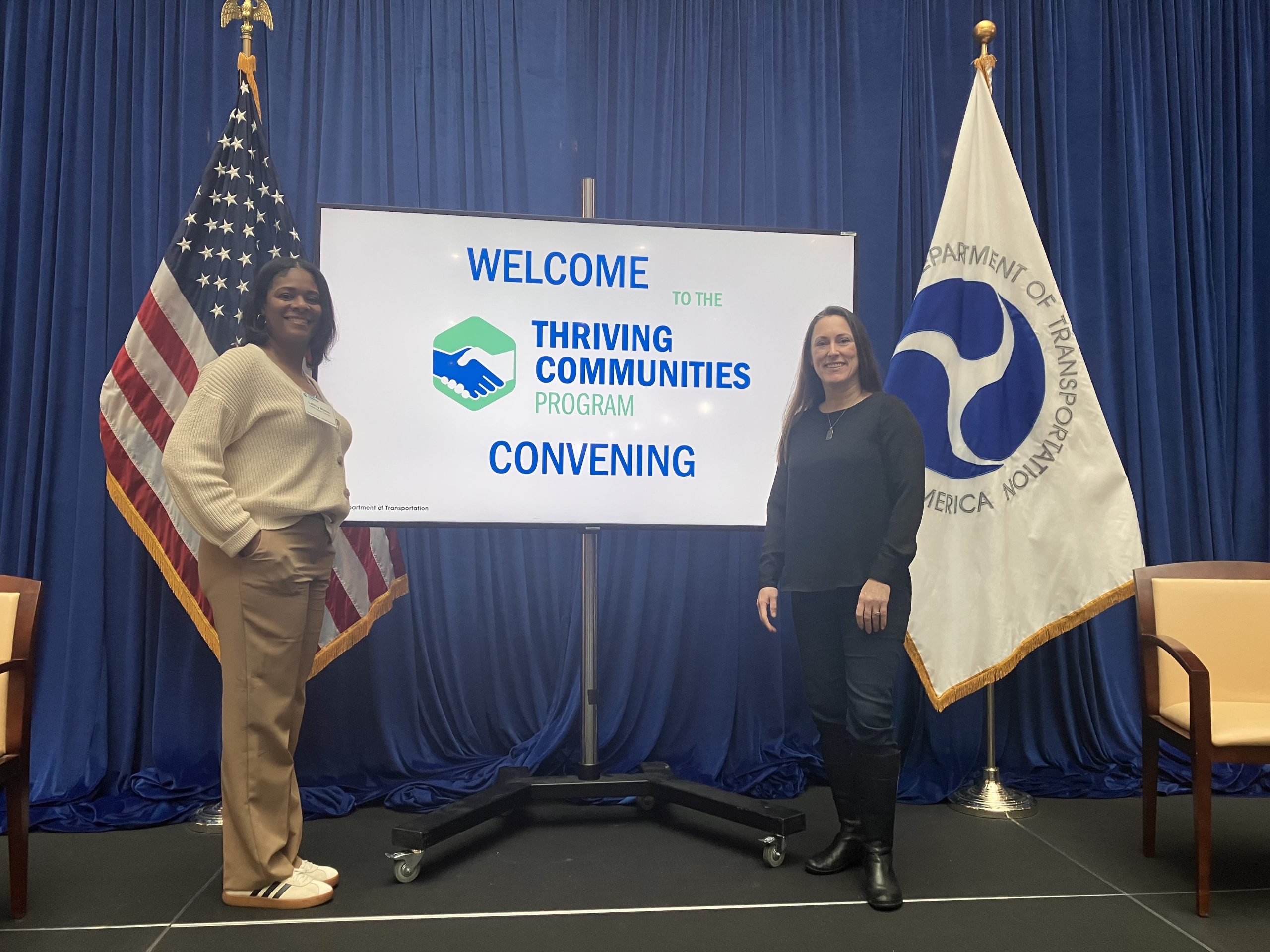 Two women stand in front of flags. A tv is between them