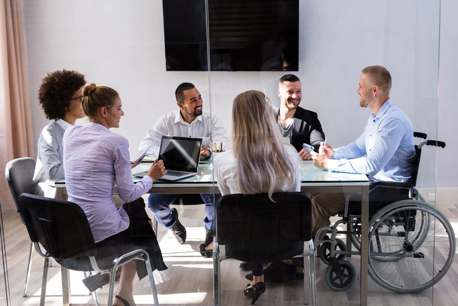 Diverse group of people, one in a wheelchair, are sitting around a table inside an office.