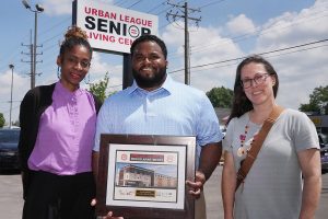 Group of three people holding a photo of a building. They are standing in front of the Urban League Senior Living Center sign