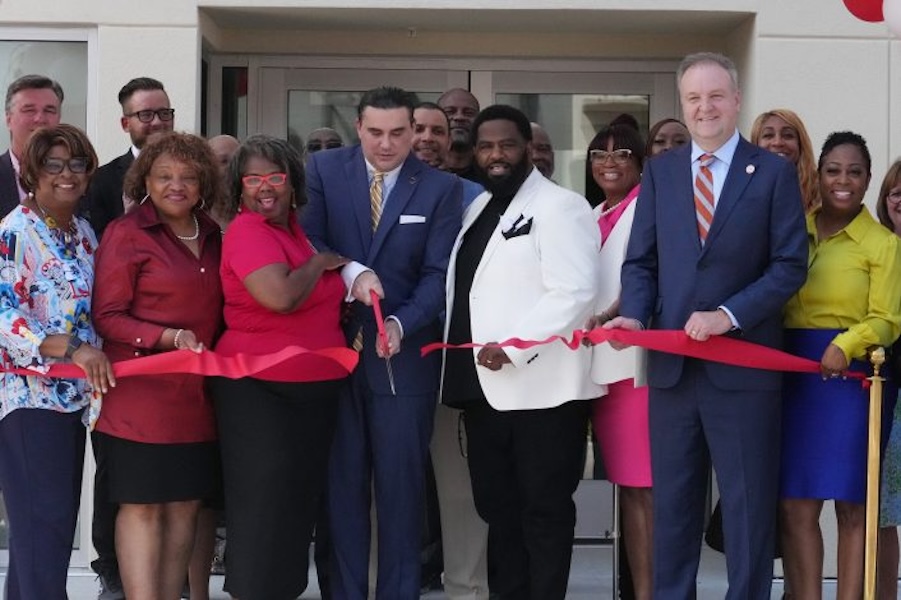 A diverse group photo of a ribbon cutting ceremony and a man in a blue suit cuts the red ribbon