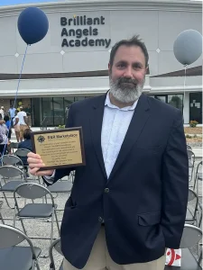 A man in a suit holds a plaque while standing in front of the Brilliant Angels Academy
