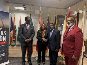 4 people stand in front of flags. All are wearing suits