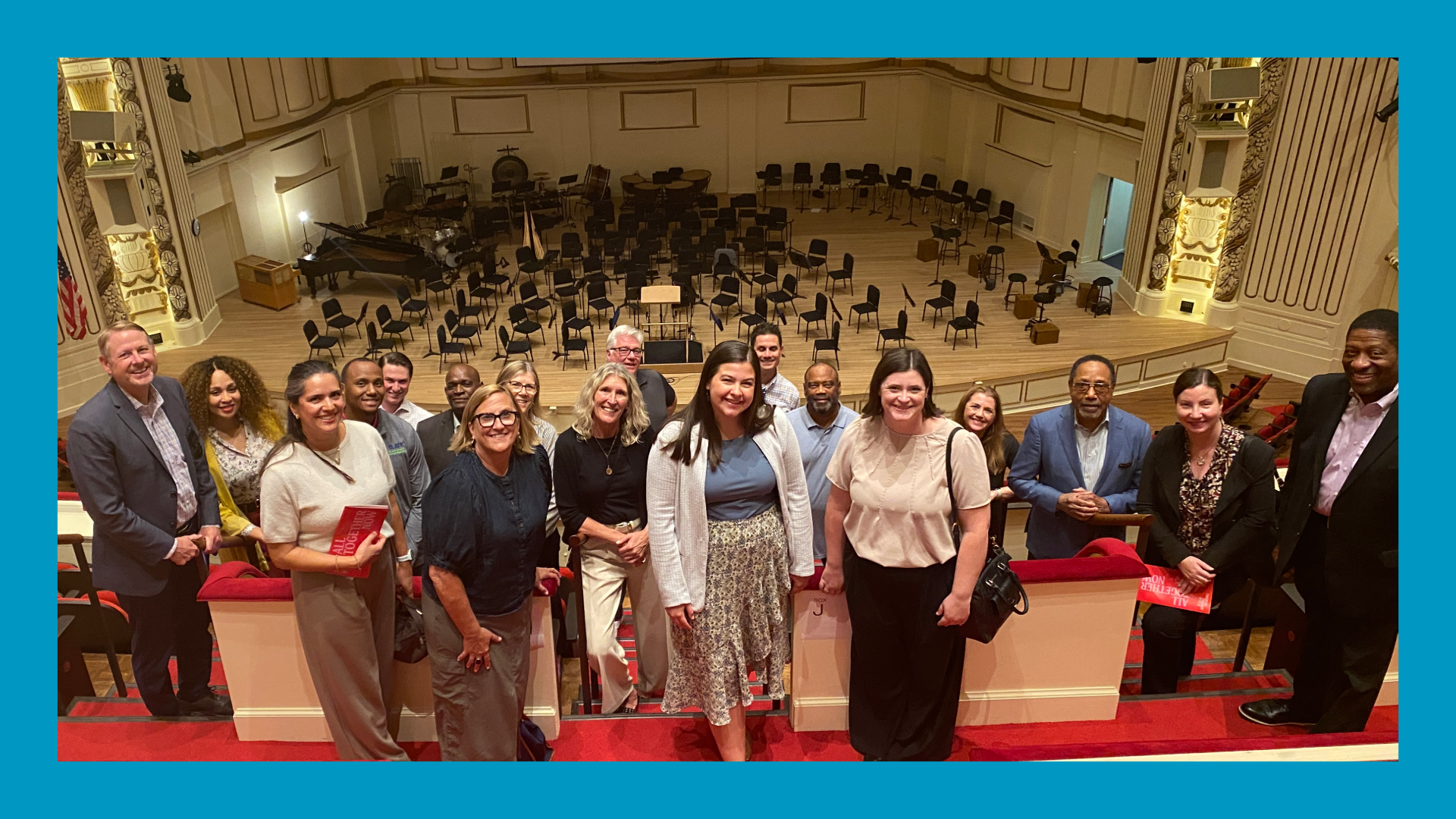 People standing in a symphony hall with the stage behind them. They are on a balcony. Blue border surrounds the image