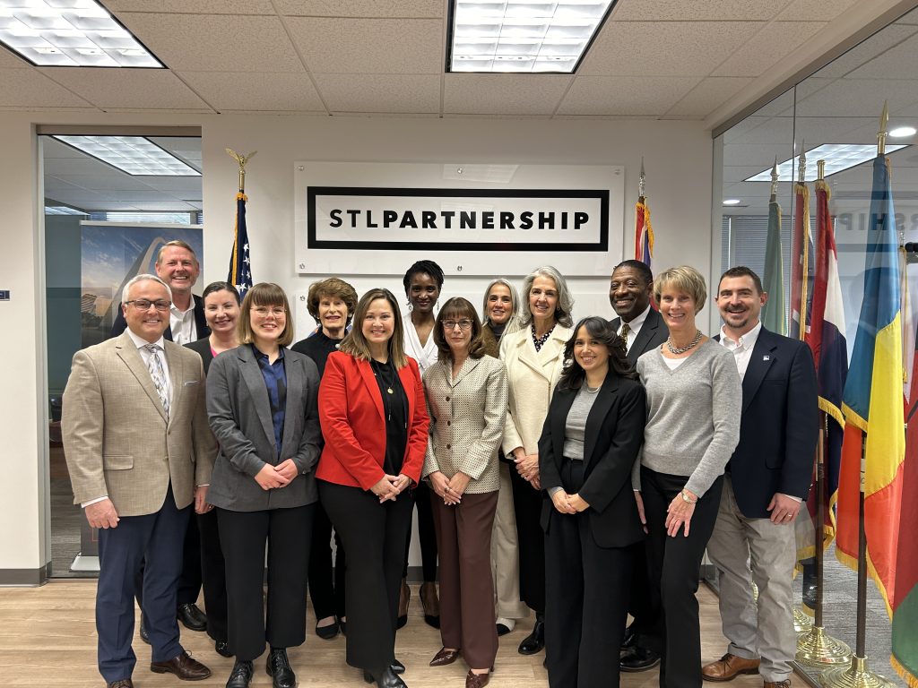 a large group of people stand posing for a picture indoors under the STL Partnership logo sign and next to flags