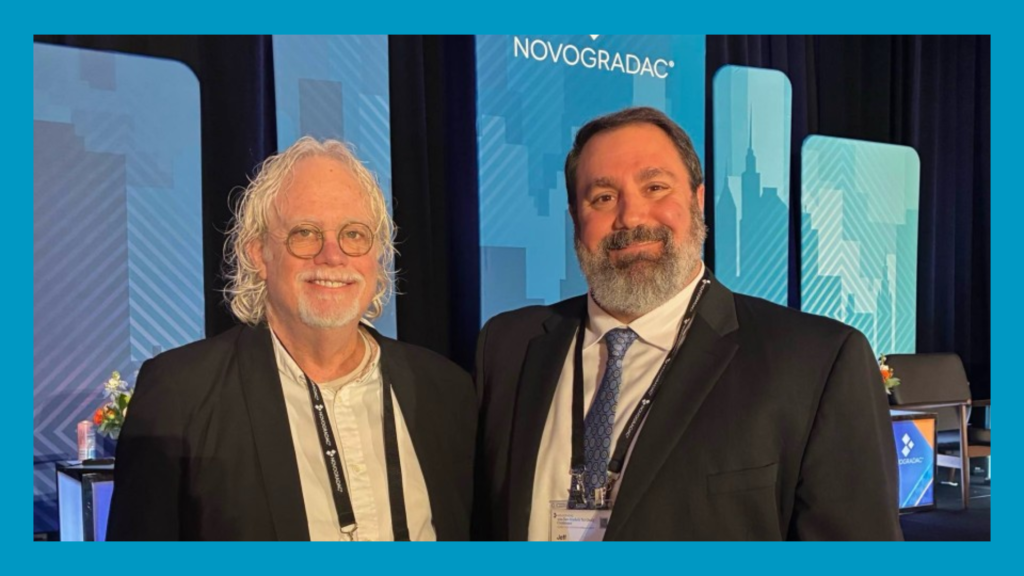 Two men stand in suits indoors smiling for camera. A blue border surrounds the picture