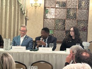 3 people sit at a table, the man in the middle speaks with a microphone