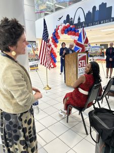 a man behind a podium smiles at Mayor spencer who stands to the side. A woman in the audience also smiles