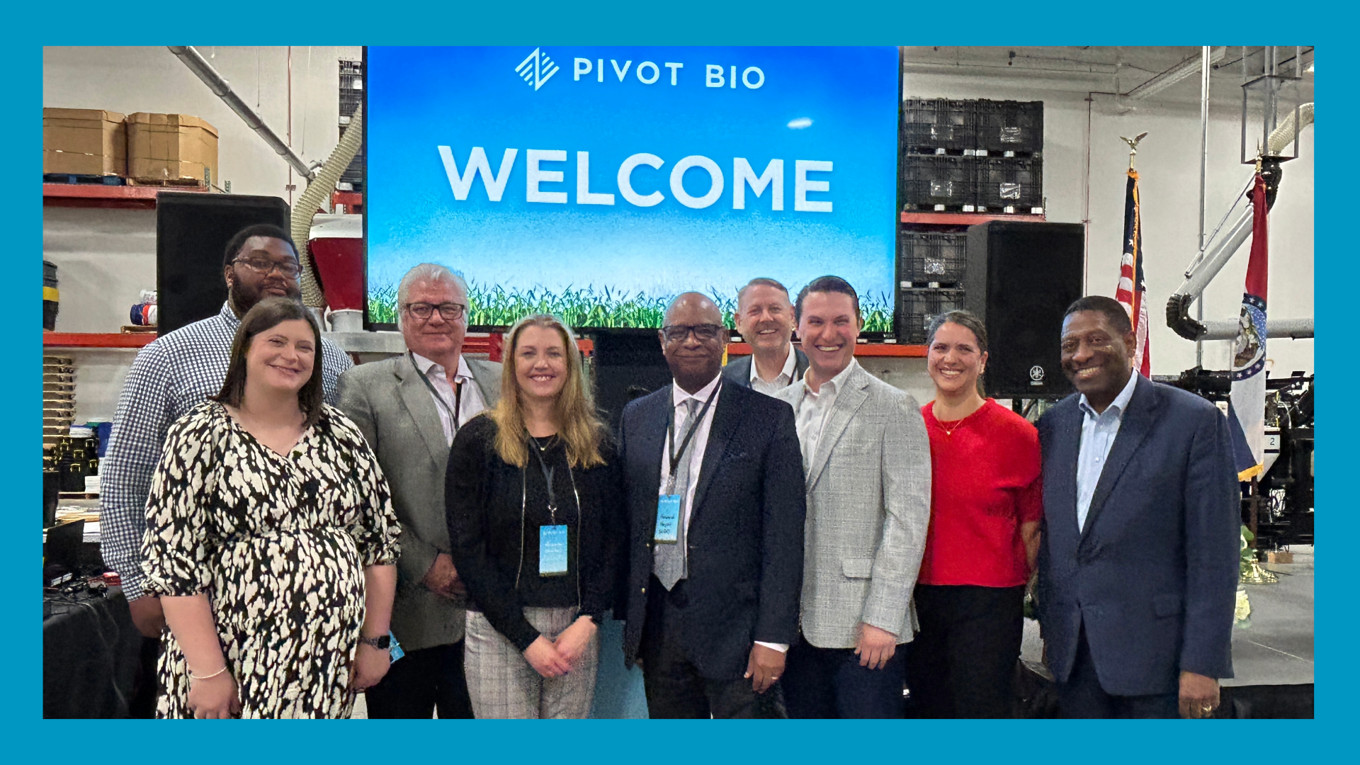 a large group of people in professional attire stand in front of a tv that says Pivot Bio - Welcome. They appear to be in a warehouse